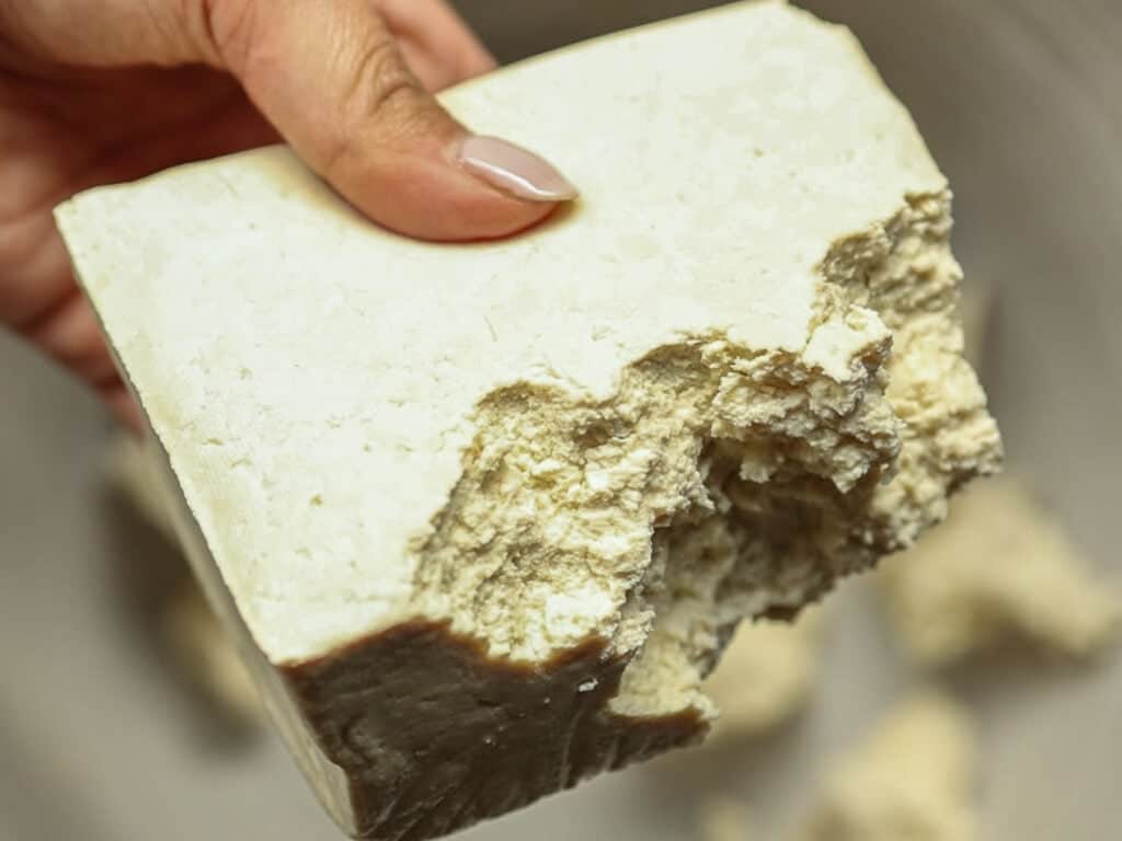 A hand holding a block of super firm tofu with pieces crumbling off. The tofu has a textured surface and a light beige color, with visible areas where chunks have broken away.